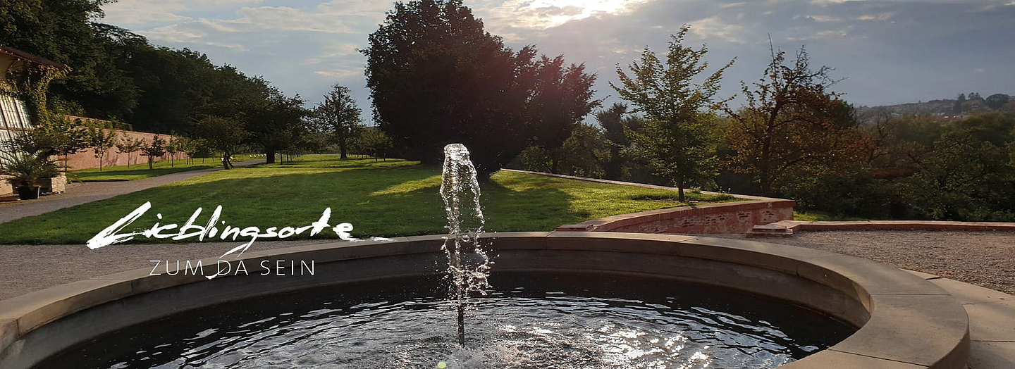Blick in einen Park mit Brunnen. "Lieblingsorte zum da sein" ist mit weißer Handschrift in das Foto geschrieben. Foto und Handschrift: Birgitta Schwansee