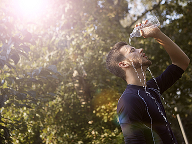 Person kühlt sich an einem heißen Sommertag mit Wasser aus einer Trinkflasche. Das Bild veranschaulicht die Auswirkungen extremer Hitze. Eine Person gießt sich im Freien Wasser aus einer Plastikflasche über Gesicht und Kopf. Im Hintergrund sind Bäume und helles Sonnenlicht zu sehen. Das Motiv steht symbolisch für hohe Temperaturen und die Belastungen durch Hitzewellen.