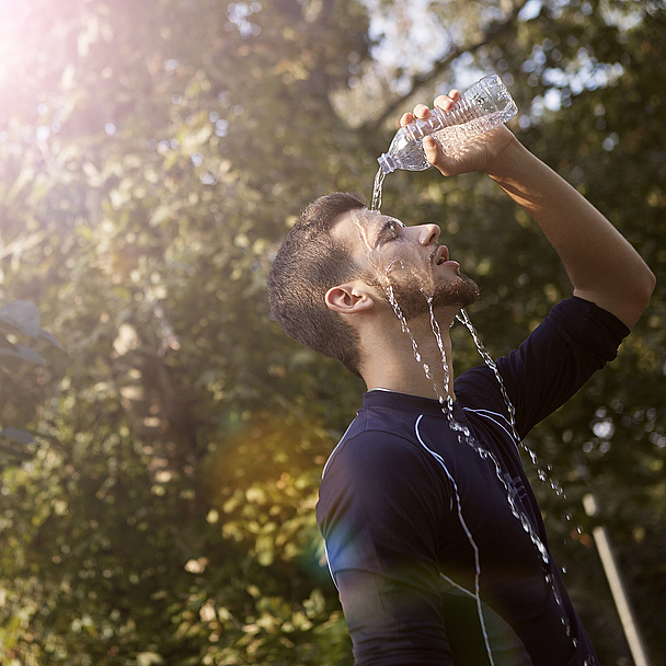 Eine Person gießt sich im Freien Wasser aus einer Plastikflasche über Gesicht und Kopf. Im Hintergrund sind Bäume und helles Sonnenlicht zu sehen. Das Motiv steht symbolisch für hohe Temperaturen und die Belastungen durch Hitzewellen.