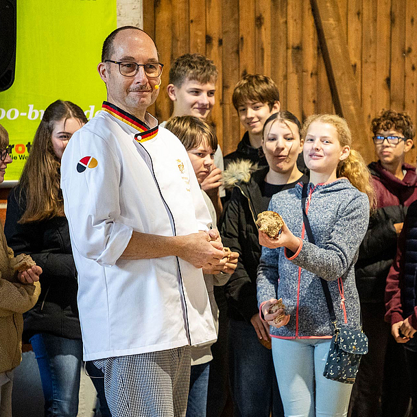 Bäckermeister Bernd Riede steht in einer Scheune und hält ein Brot. Um ihn herum stehen Konfirmandinnen und Konfirmanden aus Vellmar, die ebenfalls Brote in den Händen halten.