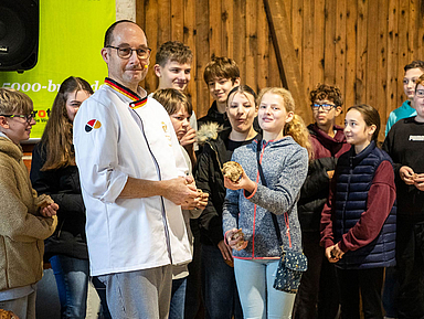 Bäckermeister Bernd Riede steht in einer Scheune und hält ein Brot. Um ihn herum stehen Konfirmandinnen und Konfirmanden aus Vellmar, die ebenfalls Brote in den Händen halten.