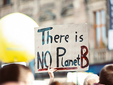 Bei einer Demonstration im Freien hält eine Person ein handgemaltes Schild hoch. Darauf steht in großen Buchstaben: „There is NO Planet B“. Im Hintergrund sind weitere Menschen, ein großes gelbes Objekt und unscharfe Gebäude zu sehen.