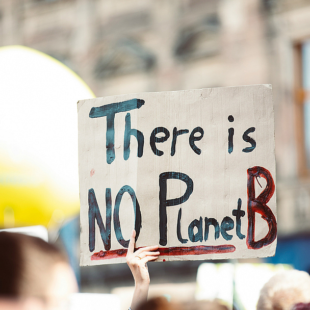 Bei einer Demonstration im Freien hält eine Person ein handgemaltes Schild hoch. Darauf steht in großen Buchstaben: „There is NO Planet B“. Im Hintergrund sind weitere Menschen, ein großes gelbes Objekt und unscharfe Gebäude zu sehen.
