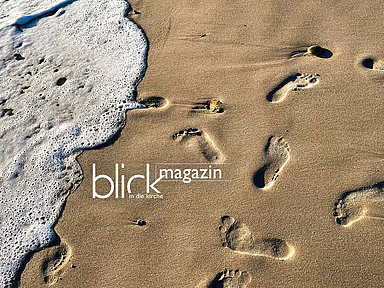 Das Bild zeigt eine Strandaufnahme aus der Nähe: Auf der rechten Seite ist trockener, heller Sand mit mehreren deutlich sichtbaren Fußspuren. Links rollt eine Welle mit weißem Schaum an den Strand und berührt einzelne Abdrücke. Kleine Steine liegen verstreut im Sand. Die Perspektive ist leicht von oben, sodass die Spuren diagonal ins Bild führen. In der Mitte links ist der Schriftzug „blick magazin“ zu sehen, darunter kleiner „in die kirche“. Die Szene wirkt ruhig und reduziert, mit Kontrast zwischen bewegtem Wasser und festem Sand.