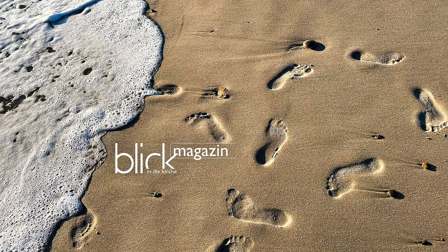 Das Bild zeigt eine Strandaufnahme aus der Nähe: Auf der rechten Seite ist trockener, heller Sand mit mehreren deutlich sichtbaren Fußspuren. Links rollt eine Welle mit weißem Schaum an den Strand und berührt einzelne Abdrücke. Kleine Steine liegen verstreut im Sand. Die Perspektive ist leicht von oben, sodass die Spuren diagonal ins Bild führen. In der Mitte links ist der Schriftzug „blick magazin“ zu sehen, darunter kleiner „in die kirche“. Die Szene wirkt ruhig und reduziert, mit Kontrast zwischen bewegtem Wasser und festem Sand.