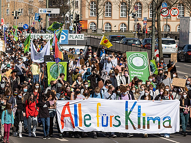 Eine große Gruppe von Menschen zieht bei einem früheren Aktionstag durch Kassel. Die Demonstrierenden marschieren auf einer Straße und tragen bunte Kleidung, Schilder und Banner. Im Vordergrund ist ein großes weißes Banner mit der Aufschrift „Alle fürs Klima“ in bunten Buchstaben zu sehen. Einige Teilnehmende tragen Masken. Im Hintergrund sind Gebäude und Straßenschilder zu erkennen
