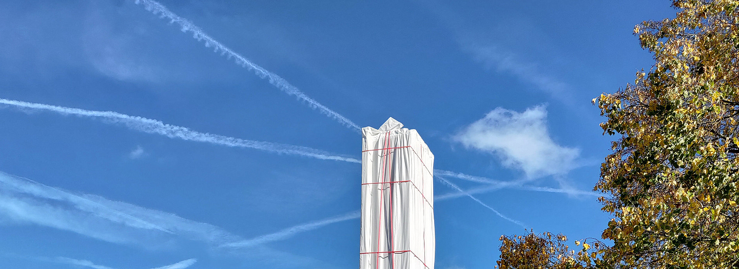 Turm in weißer Plane eingehüllt, moderne Gebäude und blauer Himmel mit Kondensstreifen. Foto (Ausschnitt): Christian Schmitt, pfarrbriefservice.de