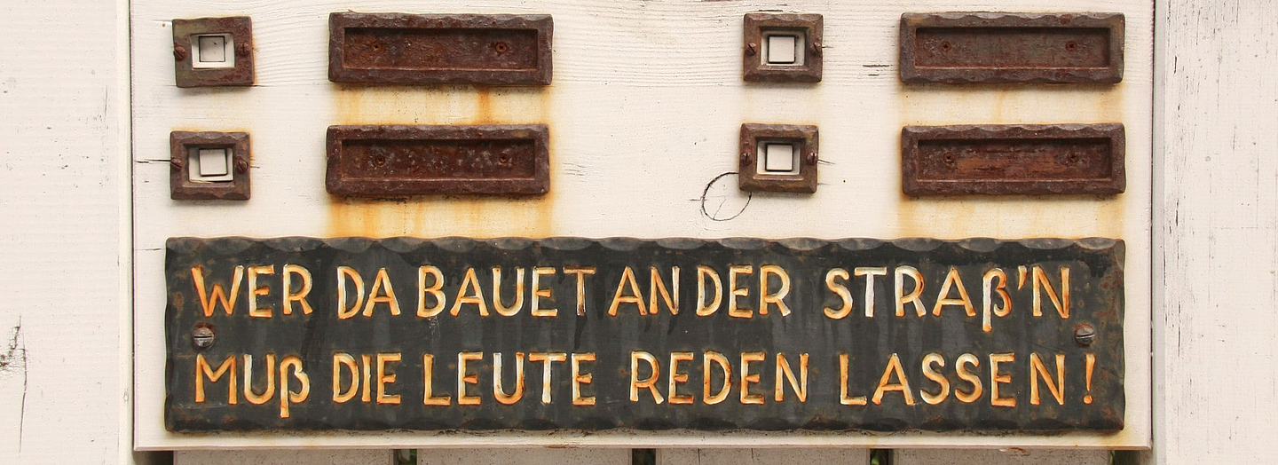 Blick auf Klingel und Klingelschilder an einem Haus mit dem Text "Wer da bauet an der Straß'n, muss die Leute reden lassen"