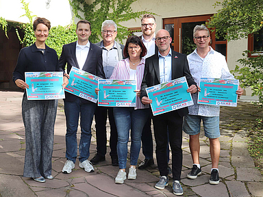 Sieben Personen stehen nebeneinander im Freien und halten jeweils ein großes, buntes Plakat in den Händen. Es handelt sich um Mitglieder des Vorbereitungsteams für den Handwerksgottesdienst. Von links nach rechts sind zu sehen: Gisa Voßeler-König, Vorsitzende des Gemeindevorstandes; Pfarrer Jan Michel Zschörnig; Peter Grohme von der EKKW; Handwerksmeisterin Elke Beyer; Marius Jung, Geschäftsführer der Kreishandwerkerschaft; sein Stellvertreter Sven Dopatka und Handwerksmeister Uwe Loth.