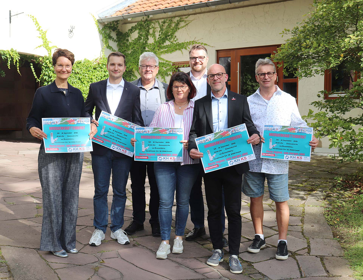 Sieben Personen stehen nebeneinander im Freien und halten jeweils ein großes, buntes Plakat in den Händen. Es handelt sich um Mitglieder des Vorbereitungsteams für den Handwerksgottesdienst. Von links nach rechts sind zu sehen: Gisa Voßeler-König, Vorsitzende des Gemeindevorstandes; Pfarrer Jan Michel Zschörnig; Peter Grohme von der EKKW; Handwerksmeisterin Elke Beyer; Marius Jung, Geschäftsführer der Kreishandwerkerschaft; sein Stellvertreter Sven Dopatka und Handwerksmeister Uwe Loth.