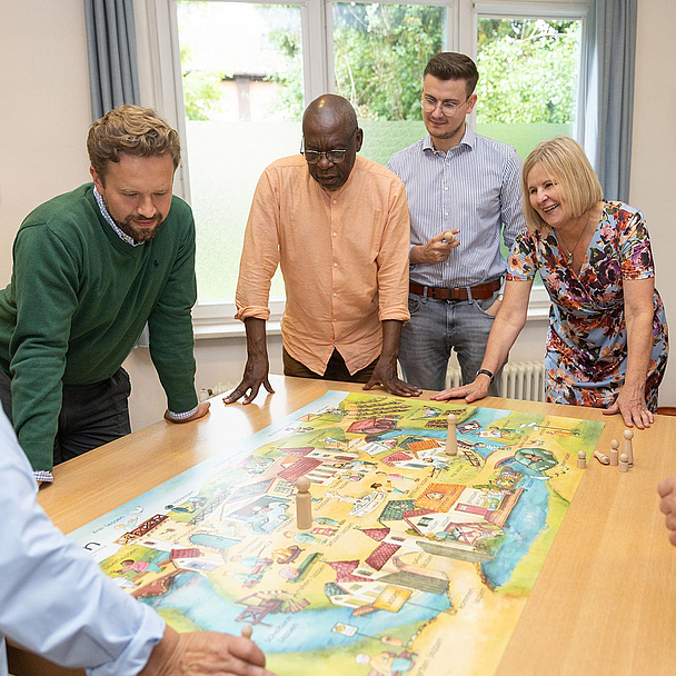 Sieben Personen stehen oder lehnen sich um einen Tisch, auf dem ein großes, buntes Spielbrett liegt. Es handelt sich um ein Planspiel zur Kirchenvorstandsarbeit. Die Gruppe wirkt konzentriert und diskutiert offenbar aktiv über das Spielgeschehen. Pfarrerin Ulrike Joachimi (vierte von rechts) nimmt ebenfalls teil. Im Hintergrund ist ein Fenster mit Blick ins Grüne zu sehen.