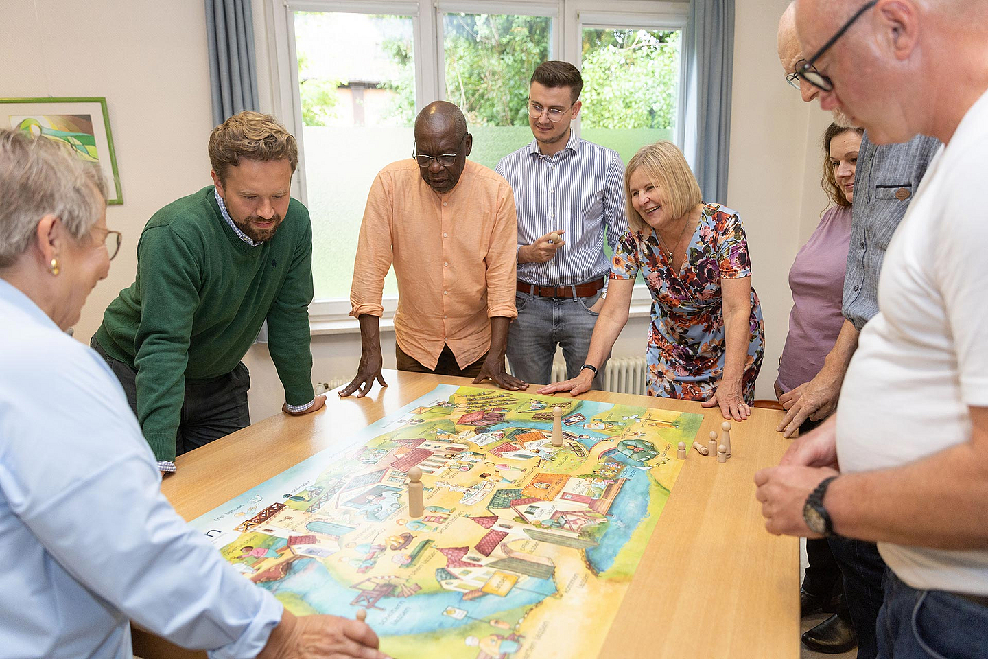 Sieben Personen stehen oder lehnen sich um einen Tisch, auf dem ein großes, buntes Spielbrett liegt. Es handelt sich um ein Planspiel zur Kirchenvorstandsarbeit. Die Gruppe wirkt konzentriert und diskutiert offenbar aktiv über das Spielgeschehen. Pfarrerin Ulrike Joachimi (vierte von rechts) nimmt ebenfalls teil. Im Hintergrund ist ein Fenster mit Blick ins Grüne zu sehen.