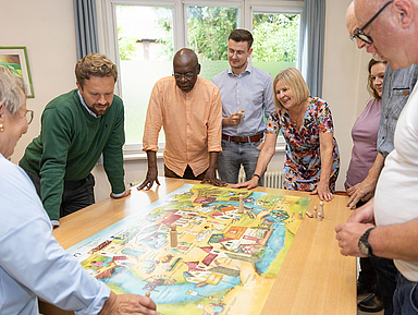 Sieben Personen stehen oder lehnen sich um einen Tisch, auf dem ein großes, buntes Spielbrett liegt. Es handelt sich um ein Planspiel zur Kirchenvorstandsarbeit. Die Gruppe wirkt konzentriert und diskutiert offenbar aktiv über das Spielgeschehen. Pfarrerin Ulrike Joachimi (vierte von rechts) nimmt ebenfalls teil. Im Hintergrund ist ein Fenster mit Blick ins Grüne zu sehen.