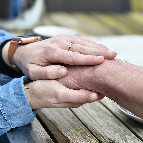 Zwei Hände liegen auf einem Holztisch. Eine Person legt beide Hände schützend und unterstützend um den Unterarm einer anderen Person. Die Szene zeigt Nähe, Unterstützung und Zuwendung in einem ruhigen, natürlichen Umfeld.