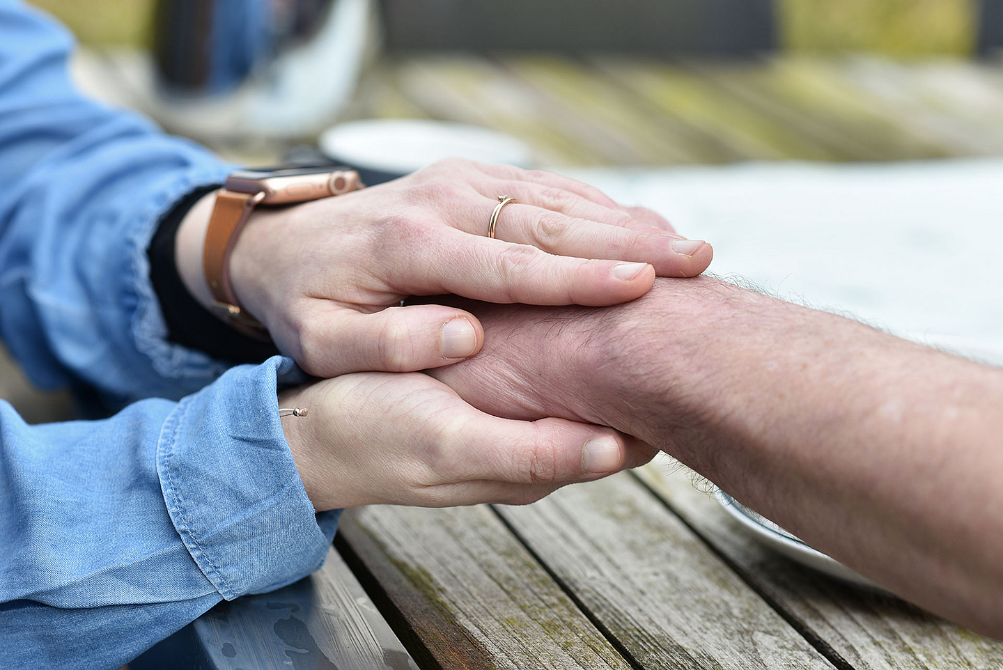 Zwei Hände liegen auf einem Holztisch. Eine Person legt beide Hände schützend und unterstützend um den Unterarm einer anderen Person. Die Szene zeigt Nähe, Unterstützung und Zuwendung in einem ruhigen, natürlichen Umfeld.