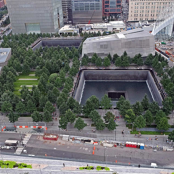 Unser Foto zeigt das 9/11-Memorial, dessen Zentrum das Mahnmal Reflecting Absence (deutsch: Spiegelung des Nichtvorhandenseins) bildet. Besucherinnen und Besucher können dort in zwei große rechteckige Hohlräume blicken, die an die Fundamente der zerstörten Zwillingstürme erinnern.