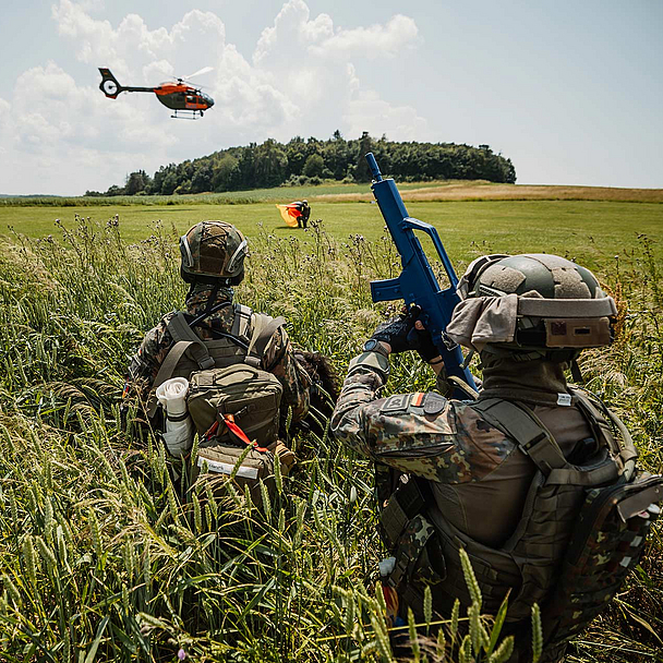 Das Bild zeigt drei Soldaten in Tarnuniform und Helmen, die im hohen Gras eines Feldes liegen. Einer trägt ein blaues Übungsgewehr. Im Hintergrund ist ein orangefarbener Hubschrauber in der Luft zu sehen, dahinter eine Baumreihe und ein weiter Himmel mit Wolken.