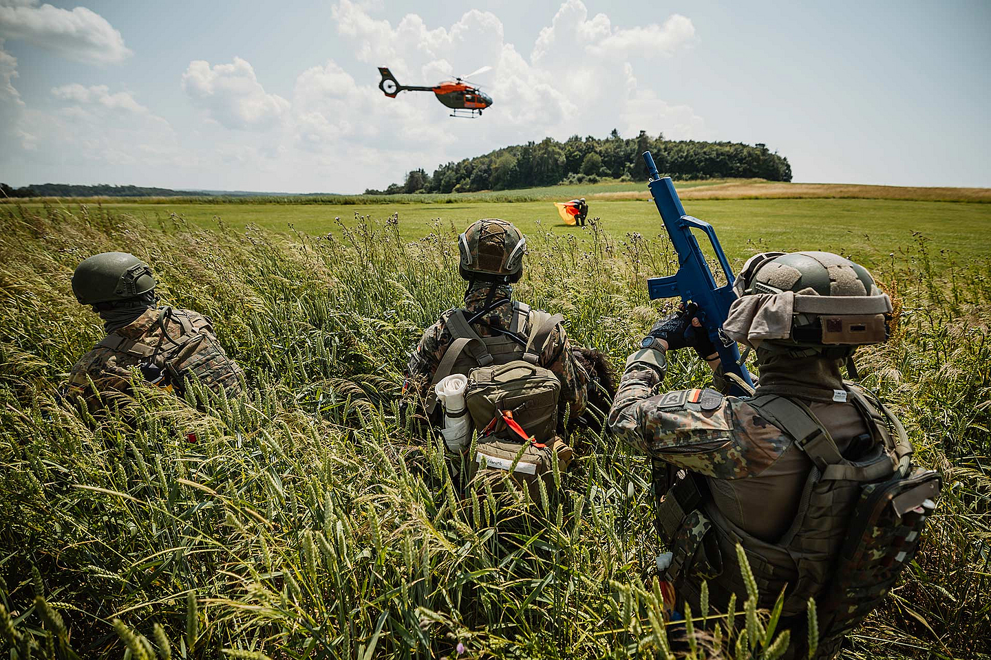Das Bild zeigt drei Soldaten in Tarnuniform und Helmen, die im hohen Gras eines Feldes liegen. Einer trägt ein blaues Übungsgewehr. Im Hintergrund ist ein orangefarbener Hubschrauber in der Luft zu sehen, dahinter eine Baumreihe und ein weiter Himmel mit Wolken.