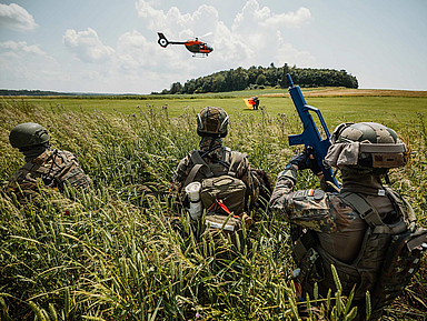 Das Bild zeigt drei Soldaten in Tarnuniform und Helmen, die im hohen Gras eines Feldes liegen. Einer trägt ein blaues Übungsgewehr. Im Hintergrund ist ein orangefarbener Hubschrauber in der Luft zu sehen, dahinter eine Baumreihe und ein weiter Himmel mit Wolken.