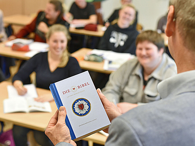 Unser Foto zeigt einen Lehrer, der mit einer Bibel in der Hand vor seiner Klasse steht.