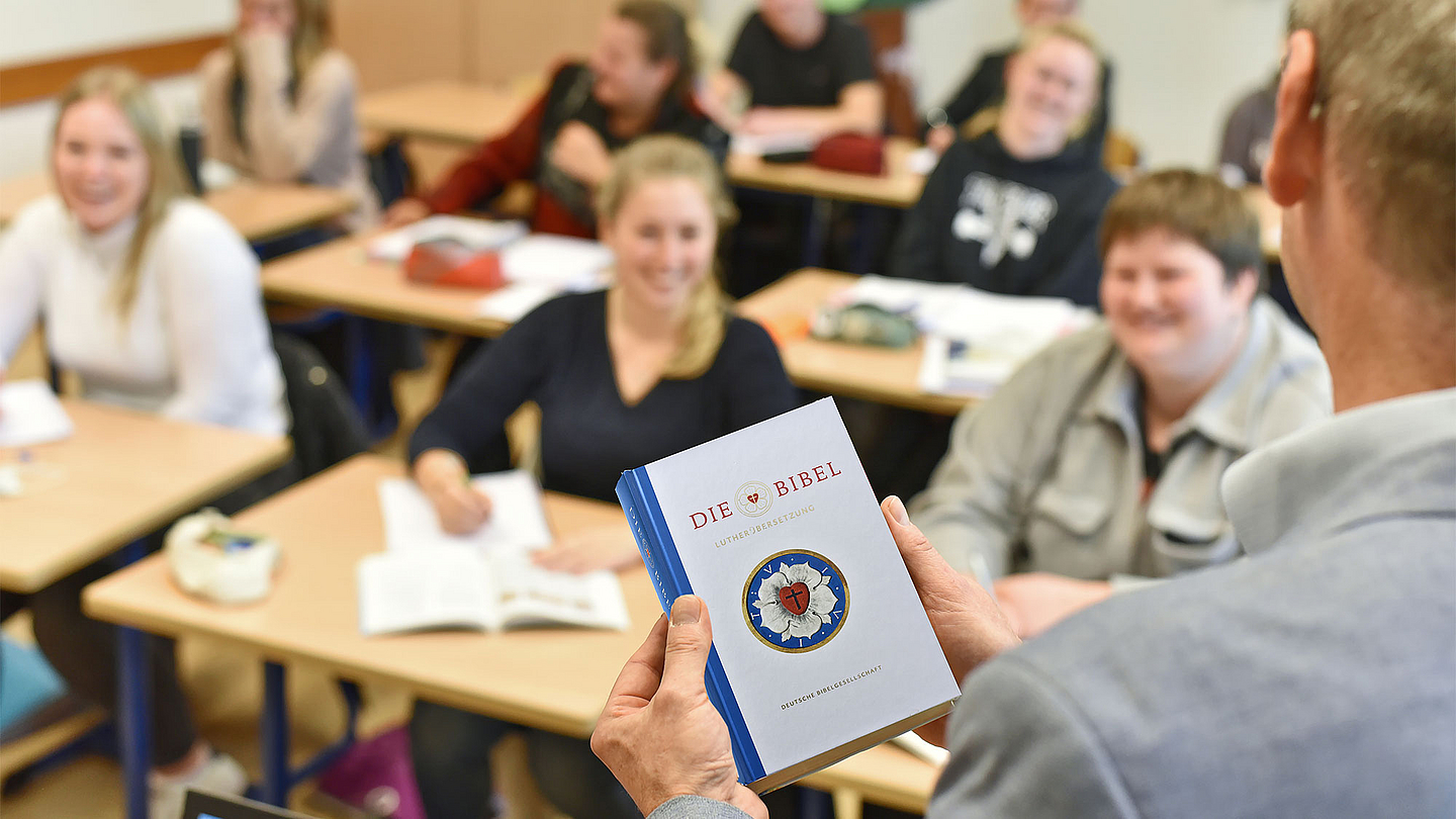 Unser Foto zeigt einen Lehrer, der mit einer Bibel in der Hand vor seiner Klasse steht.