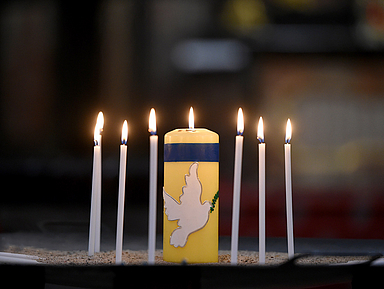 Das Bild zeigt Kerzen auf einem Altar. Darunter eine gelb-blaue Kerze mit einer Friedenstaube.