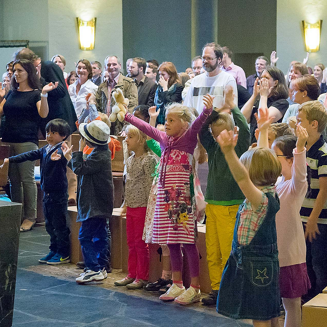Kinder stehen mit erhobenen Armen in der ersten Reihe eines gut besuchten Gottesdienstes, Erwachsene sitzen und stehen dahinter in einem hellen Kirchenraum mit Holzbänken und hoher Decke. Auf dem Boden liegen Kartons.