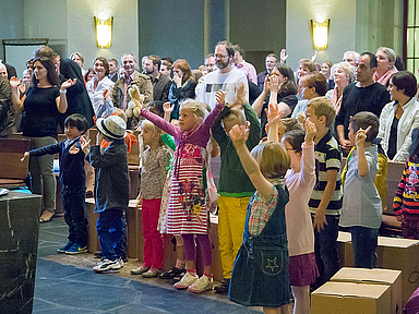 Kinder stehen mit erhobenen Armen in der ersten Reihe eines gut besuchten Gottesdienstes, Erwachsene sitzen und stehen dahinter in einem hellen Kirchenraum mit Holzbänken und hoher Decke. Auf dem Boden liegen Kartons.