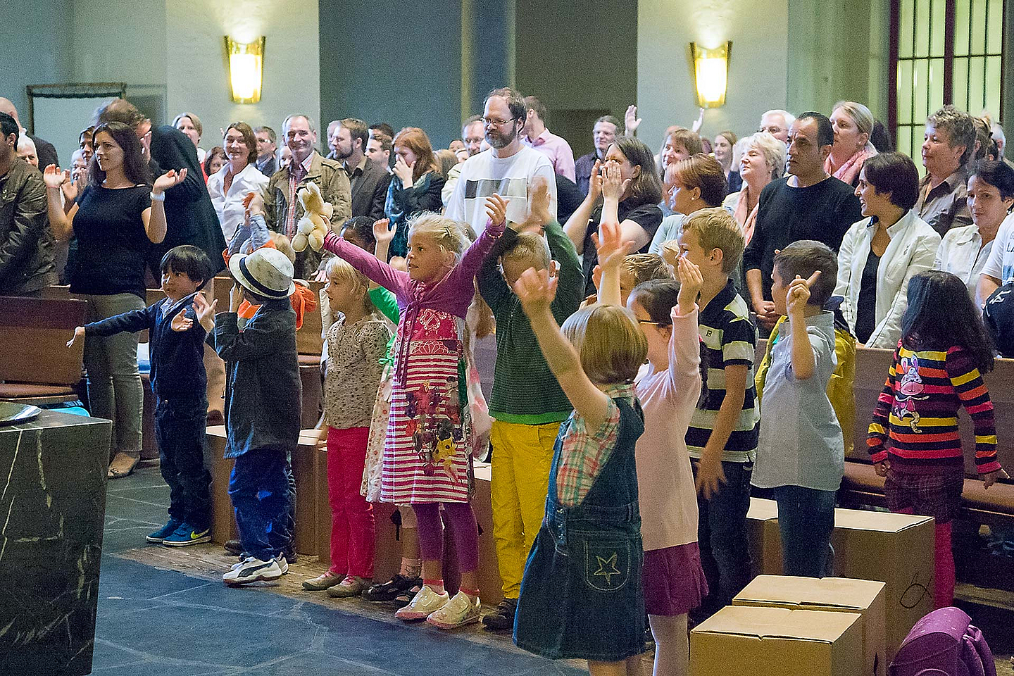 Kinder stehen mit erhobenen Armen in der ersten Reihe eines gut besuchten Gottesdienstes, Erwachsene sitzen und stehen dahinter in einem hellen Kirchenraum mit Holzbänken und hoher Decke. Auf dem Boden liegen Kartons.