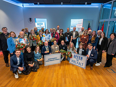 Das Bild zeigt eine Gruppe von etwa 30 Personen, die in einem Raum mit Holzboden und blauer Beleuchtung stehen. Einige Personen halten Blumensträuße und Schilder mit den Aufschriften „Offen für Vielfalt“ und „Demokratie-Verstärker*innen“. Im Hintergrund sind weitere Schilder zu sehen. In ganz links steht Kassels Regierungspräsident Mark Weinmeister, in der ersten Reihe rechts Christoph Lübcke, Sohn des ermordeten Kasseler Regierungspräsidenten Walter Lübcke.