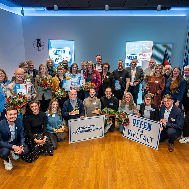 Das Bild zeigt eine Gruppe von etwa 30 Personen, die in einem Raum mit Holzboden und blauer Beleuchtung stehen. Einige Personen halten Blumensträuße und Schilder mit den Aufschriften „Offen für Vielfalt“ und „Demokratie-Verstärker*innen“. Im Hintergrund sind weitere Schilder zu sehen. In ganz links steht Kassels Regierungspräsident Mark Weinmeister, in der ersten Reihe rechts Christoph Lübcke, Sohn des ermordeten Kasseler Regierungspräsidenten Walter Lübcke.