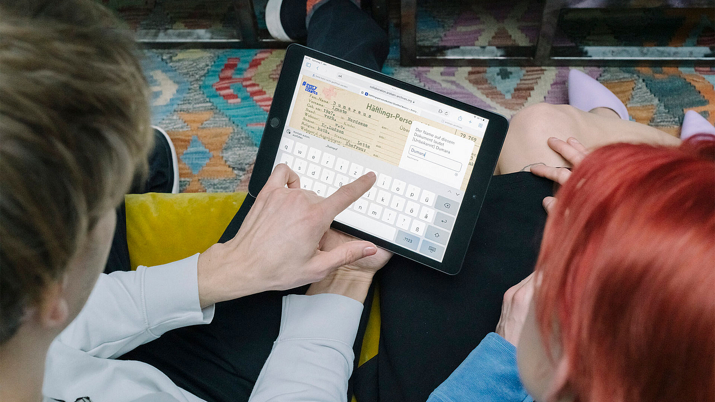 Das Bild zeigt einen Mann, der ein Tablet in der Hand hält und darauf zeigt. Darauf zu sehen ist eine Häftlingsakte. Neben ihm sitzt eine Frau. 