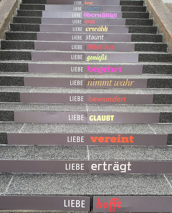 Steinerne Treppenstufen, auf deren Vorderseiten Schilder mit Textbotschaften angebracht sind. Jede Zeile beginnt mit dem Wort „LIEBE“, gefolgt von einem Verb in unterschiedlichen Farben und Schriftarten. Foto: Catrin Lerch, pfarrbriefservice.de