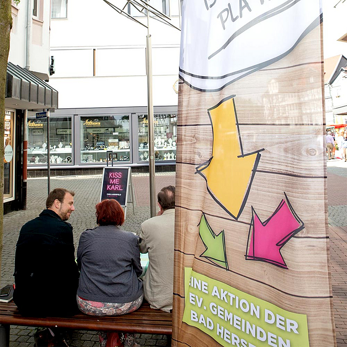 Pfarrer Jaeger (l.) im Gespräch mit zwei Gästen auf der Bank. Im Vordergrund ist die Fahne zu sehen, die auf das Angebot weithin sichbar aufmerksam macht. Pfarrer Jaeger (l.) im Gespräch mit zwei Gästen auf der Bank. Im Vordergrund ist die Fahne zu sehen, die auf das Angebot weithin sichbar aufmerksam macht.