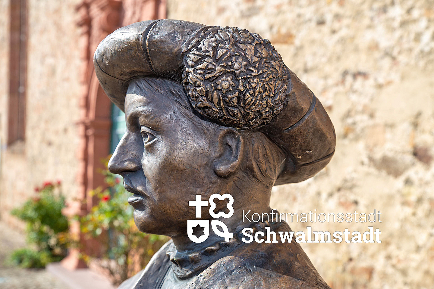 Zu sehen ist eine Statue von Landgraf Philipp I. von Hessen mit einem reich verzierten Hut. Die Figur steht auf dem Paradeplatz in Ziegenhain vor einer alten Sandsteinmauer und erinnert an die Einführung der Konfirmation durch Philipp und Martin Bucer.