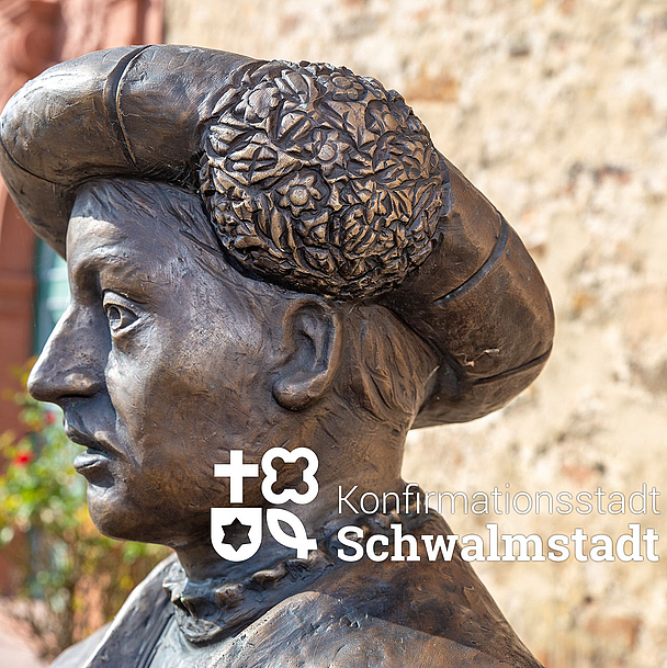 Zu sehen ist eine Statue von Landgraf Philipp I. von Hessen mit einem reich verzierten Hut. Die Figur steht auf dem Paradeplatz in Ziegenhain vor einer alten Sandsteinmauer und erinnert an die Einführung der Konfirmation durch Philipp und Martin Bucer.