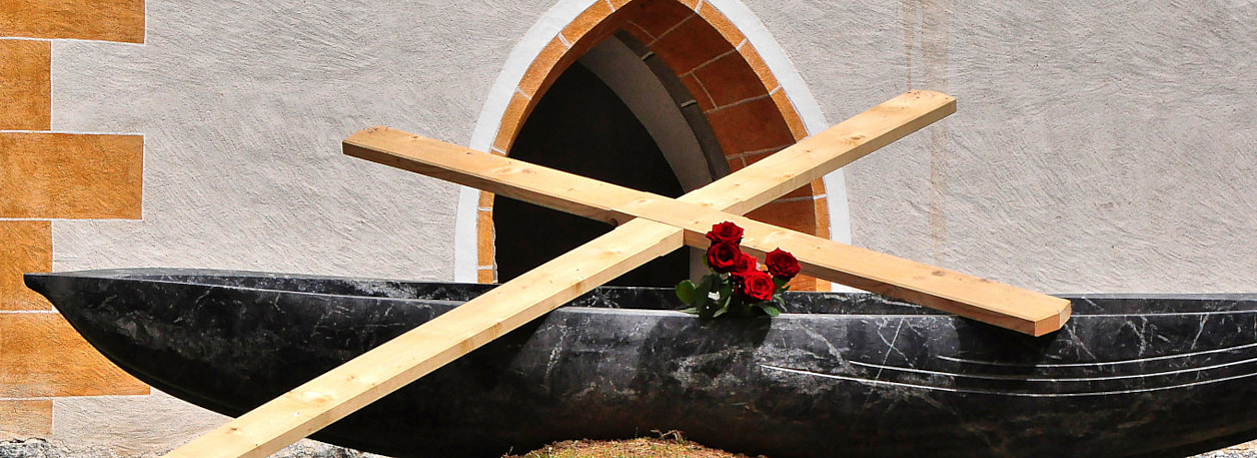 Auf einem schwarzen Boot liegt ein großes Holzkreuz und ein paar Rosen. Im Hintergund ist der Eingang zu einem Kirchengebäude zu sehen.