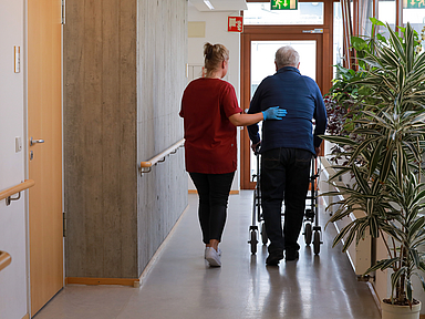 Eine Pflegekraft begleitet einen Mann mit Rollator über einen Flur.