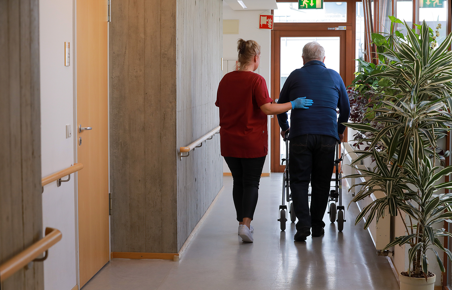 Eine Pflegekraft begleitet einen Mann mit Rollator über einen Flur.