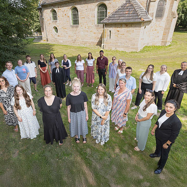 Die Gruppe von 23 Personen steht in Hofgeismar vor der Brunnenkirche, einer historischen Steinkirche mit Bogenfenstern; zwei Personen tragen schwarze Roben (Talar). Die Gruppe steht im Halbkreis auf einer Rasenfläche, im Hintergrund sind Bäume zu sehen.