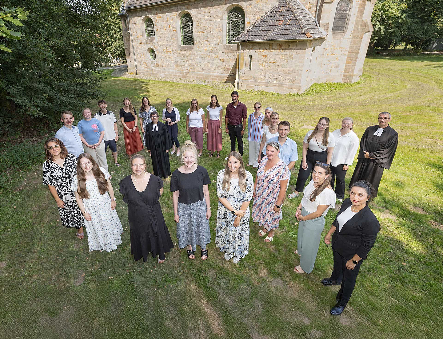 Die Gruppe von 23 Personen steht in Hofgeismar vor der Brunnenkirche, einer historischen Steinkirche mit Bogenfenstern; zwei Personen tragen schwarze Roben (Talar). Die Gruppe steht im Halbkreis auf einer Rasenfläche, im Hintergrund sind Bäume zu sehen.