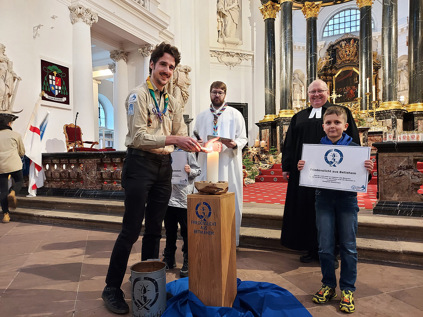 Friedenslicht-Träger 2025 Fabian Stork (DPSG, Stamm Wächtersbach) entzündet eine Kerze mit dem Friedenslicht aus Bethlehem. Im Hintergrund zu sehen sind DPSG-Diözesankurat Patrick Jestädt (l.) und Pfarrer Jörg Scheer von der evangelischen Christuskirche in Fulda. 