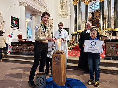 Friedenslicht-Träger 2025 Fabian Stork (DPSG, Stamm Wächtersbach) entzündet eine Kerze mit dem Friedenslicht aus Bethlehem. Im Hintergrund zu sehen sind DPSG-Diözesankurat Patrick Jestädt (l.) und Pfarrer Jörg Scheer von der evangelischen Christuskirche in Fulda. 