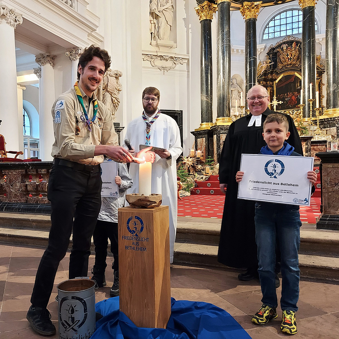 Friedenslicht-Träger 2025 Fabian Stork (DPSG, Stamm Wächtersbach) entzündet eine Kerze mit dem Friedenslicht aus Bethlehem. Im Hintergrund zu sehen sind DPSG-Diözesankurat Patrick Jestädt (l.) und Pfarrer Jörg Scheer von der evangelischen Christuskirche in Fulda. 