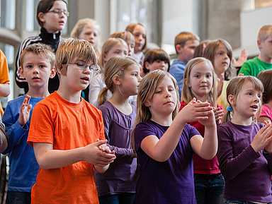 Ein Kinder eines Chores stehen dicht beieinander und klatschen rhythmisch. Sie tragen bunte Kleidung in Orange, Blau, Lila und Grün. Das Foto wurde in der Kasseler Martinskirche aufgenommen.