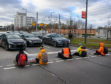 Das Foto zeigt eine Straßenblockade der Protestgruppe „Letzte Generation“ in Berlin im Jahr 2022. Fünf Personen sitzen auf der Fahrbahn und haben sich aus Protest gegen mangelnden Klimaschutz an die Straße geklebt. Sie tragen auffällige Jacken in Orange und Gelb sowie Rucksäcke. Einige halten Schilder oder Transparente. Im Hintergrund stehen Autos, die durch die Aktion gestoppt wurden. Zu sehen sind außerdem städtische Elemente wie Gebäude, Straßenbahnschienen und Verkehrsschilder.