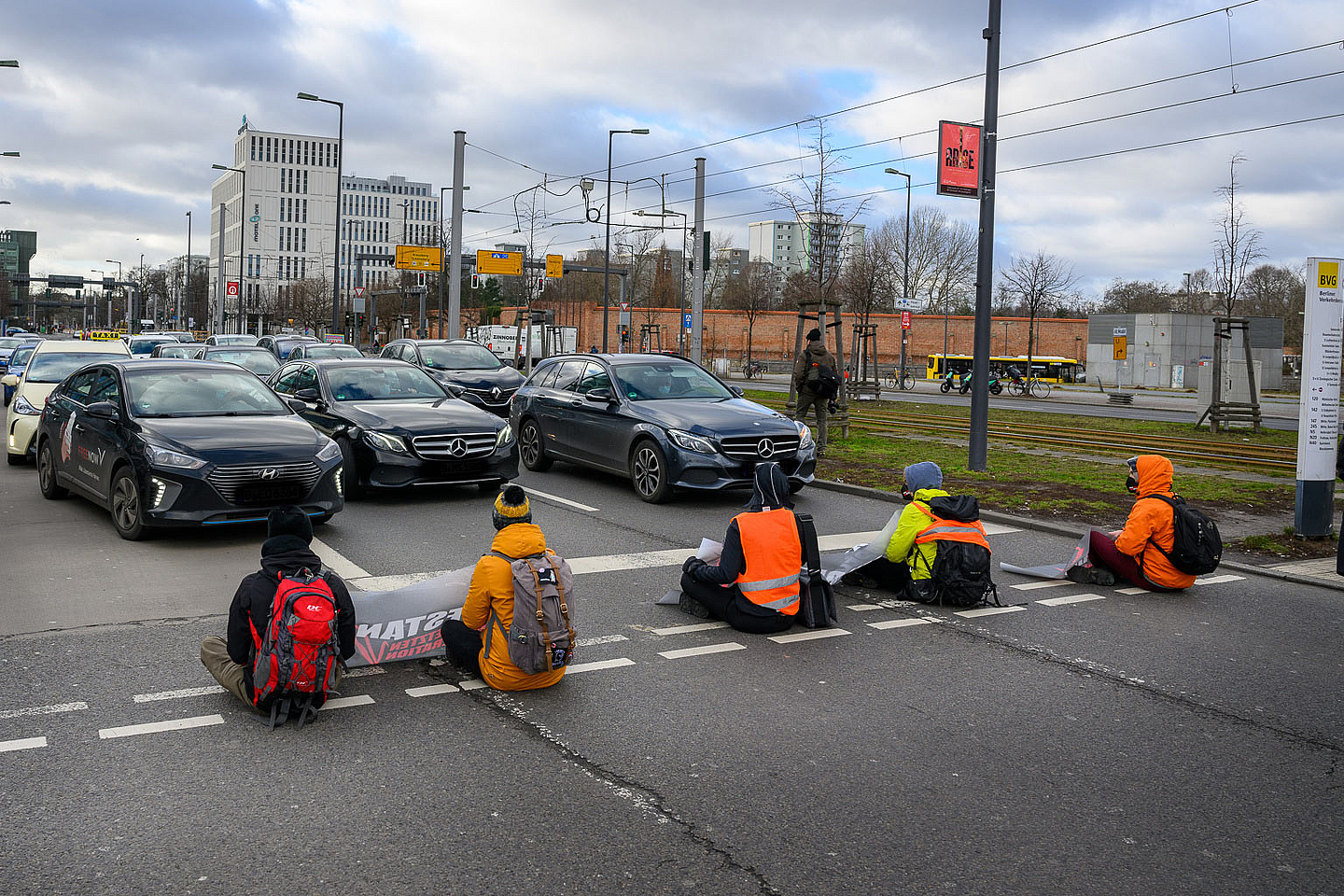 Das Foto zeigt eine Straßenblockade der Protestgruppe „Letzte Generation“ in Berlin im Jahr 2022. Fünf Personen sitzen auf der Fahrbahn und haben sich aus Protest gegen mangelnden Klimaschutz an die Straße geklebt. Sie tragen auffällige Jacken in Orange und Gelb sowie Rucksäcke. Einige halten Schilder oder Transparente. Im Hintergrund stehen Autos, die durch die Aktion gestoppt wurden. Zu sehen sind außerdem städtische Elemente wie Gebäude, Straßenbahnschienen und Verkehrsschilder.