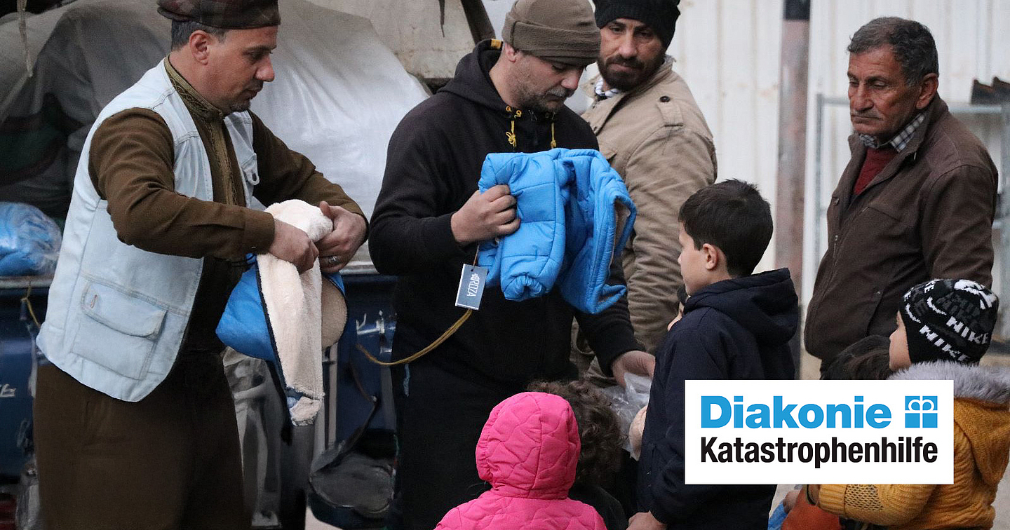 Eine Gruppe von Personen verteilt Kleidung an Kinder und Erwachsene im Freien; eine Person überreicht eine blaue Jacke, eine andere hält eine Decke. Das Logo „Diakonie Katastrophenhilfe“ ist unten rechts sichtbar.