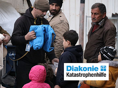 Eine Gruppe von Personen verteilt Kleidung an Kinder und Erwachsene im Freien; eine Person überreicht eine blaue Jacke, eine andere hält eine Decke. Das Logo „Diakonie Katastrophenhilfe“ ist unten rechts sichtbar.