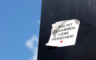 Zettel mit Aufschrift „Haltet zusammen, liebe Menschen!“ an schwarzer Wand, blauer Himmel. Foto: Peter Weidemann, pfarrbriefservice.de